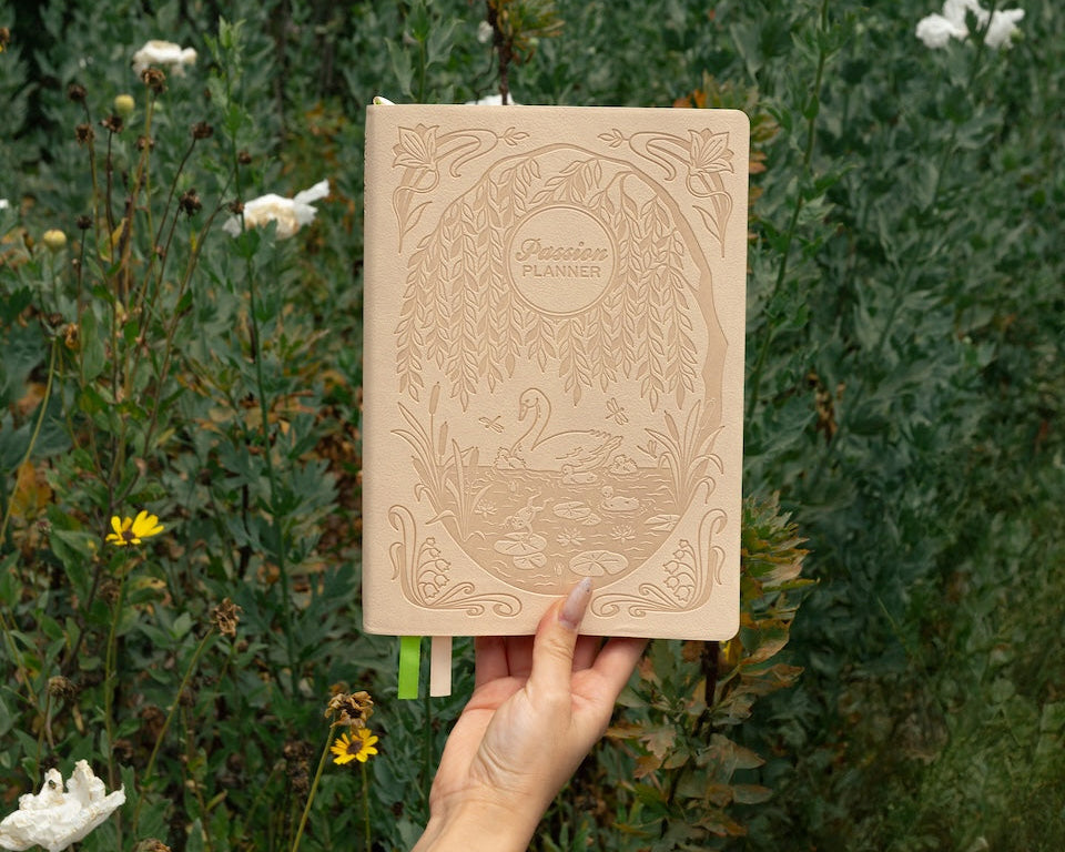 Hand holding a beige notebook with decorative swan cover against a natural background of greenery and white flowers