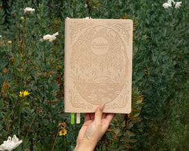 Hand holding a beige notebook with decorative swan cover against a natural background of greenery and white flowers