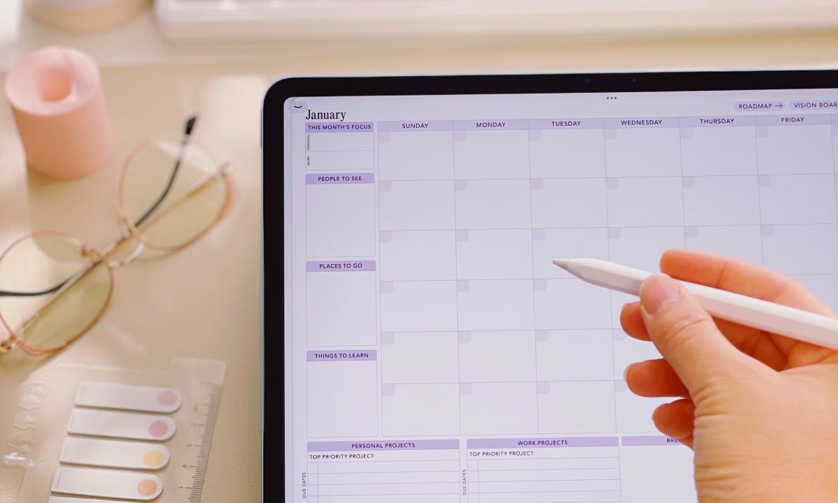 Person using a digital calendar on a tablet with a hand holding a stylus, surrounded by office items.
