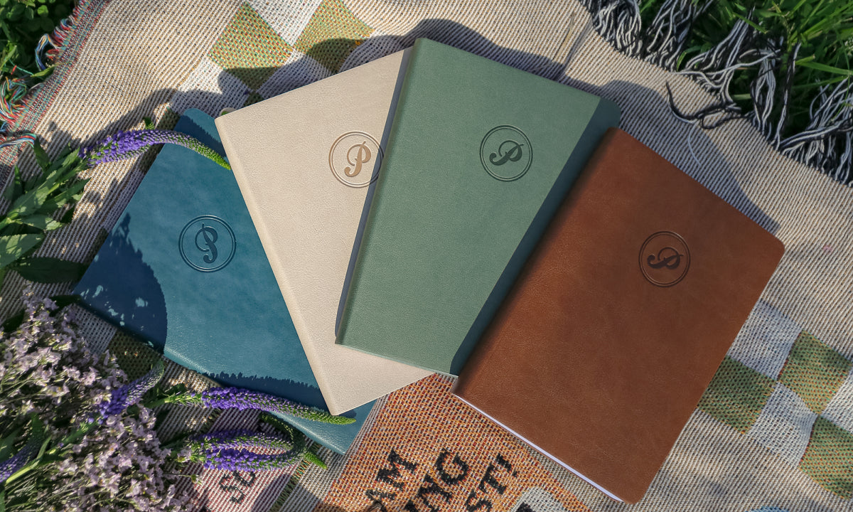 Four notebooks in different colors with a passion planner logo on a textured surface with plants.