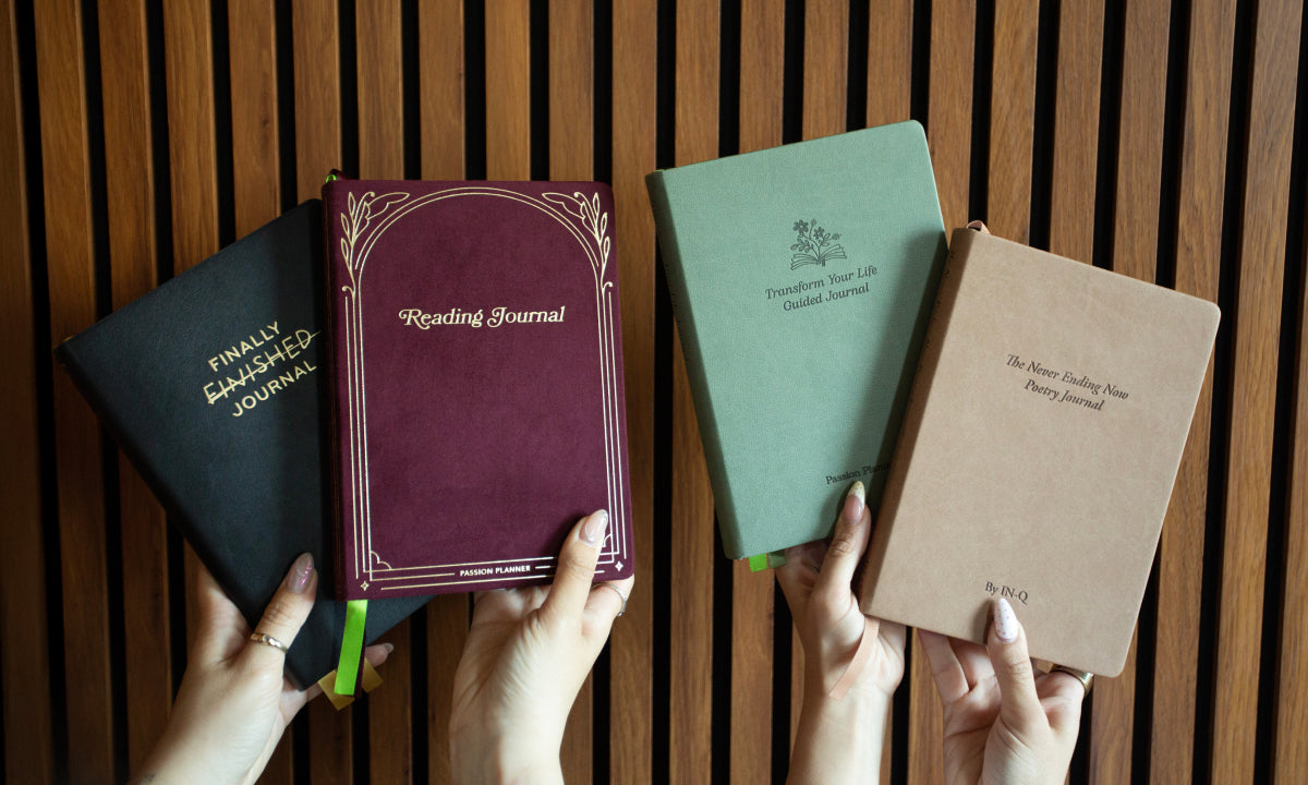 Four journals of different colors held by hands against a wooden background