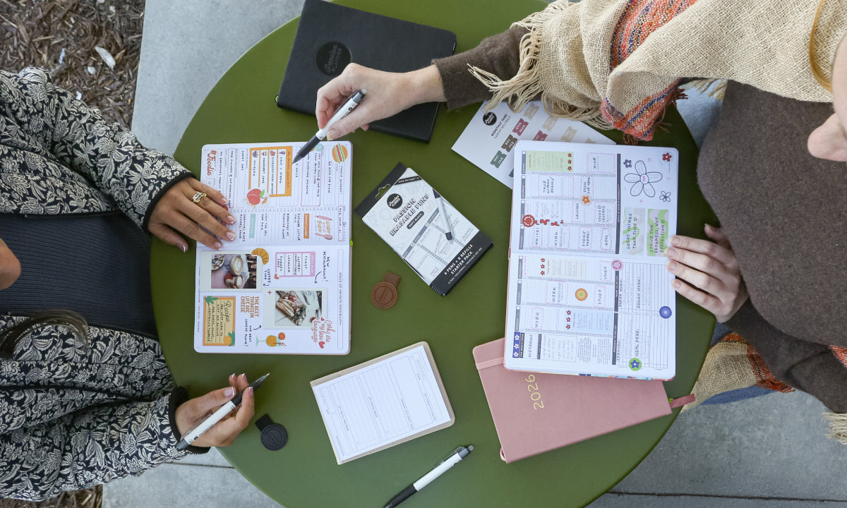 Two people sitting at a round green table with papers, notebooks, and pens.