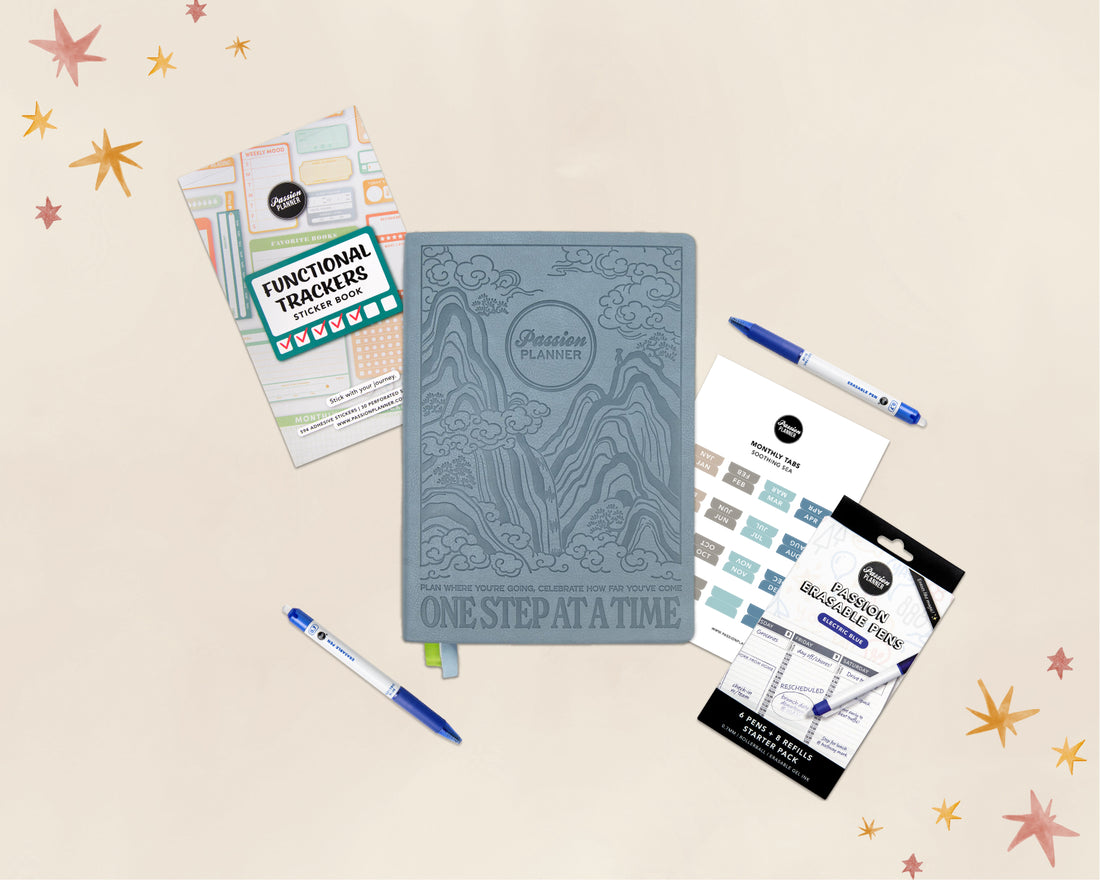 A styled flat-lay of a planner set on a warm cream background with decorative star graphics. The main item is a slate blue embossed &