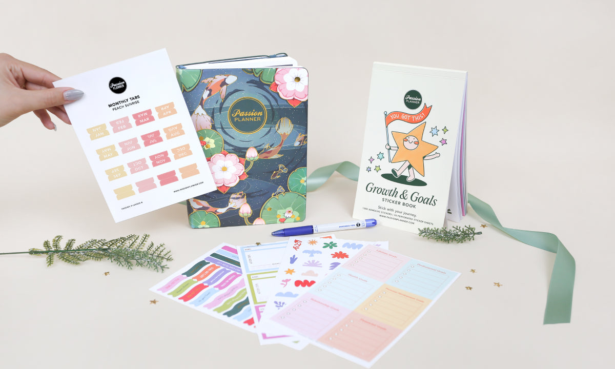 Collection of planners and stationery items on a light surface with a hand holding a monthly tabs sticker sheet.