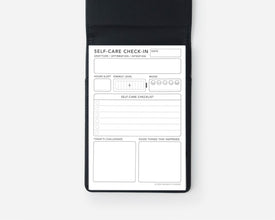 A black self-care checklist notepad shown open against a light gray background, featuring a white insert with fields for date, self-care check-in, sleep, energy level, mood, challenges, and good things that happened.