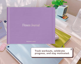 A purple Fitness Journal by Passion Planner is displayed on a desk with a tablet, notebook, and small retro TV prop. A text bubble reads: 'Track workouts, celebrate progress, and stay motivated.'.