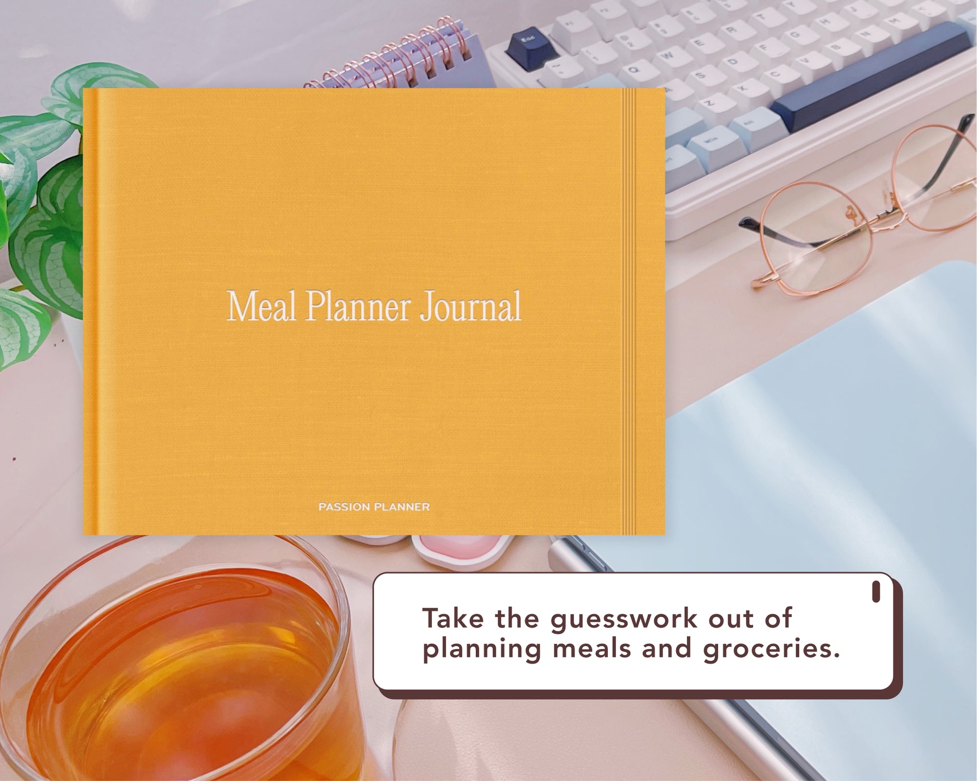 A yellow-orange digital journal titled 'Meal Planner Journal' displayed on a desk with a keyboard, glasses, tea, and a small plant. A promotional badge at the bottom reads, 'Take the guesswork out of planning meals and groceries.