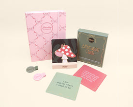 Collection of planner accessories including a pink planner, card with mushroom design, and affirmation cards on a light beige background.