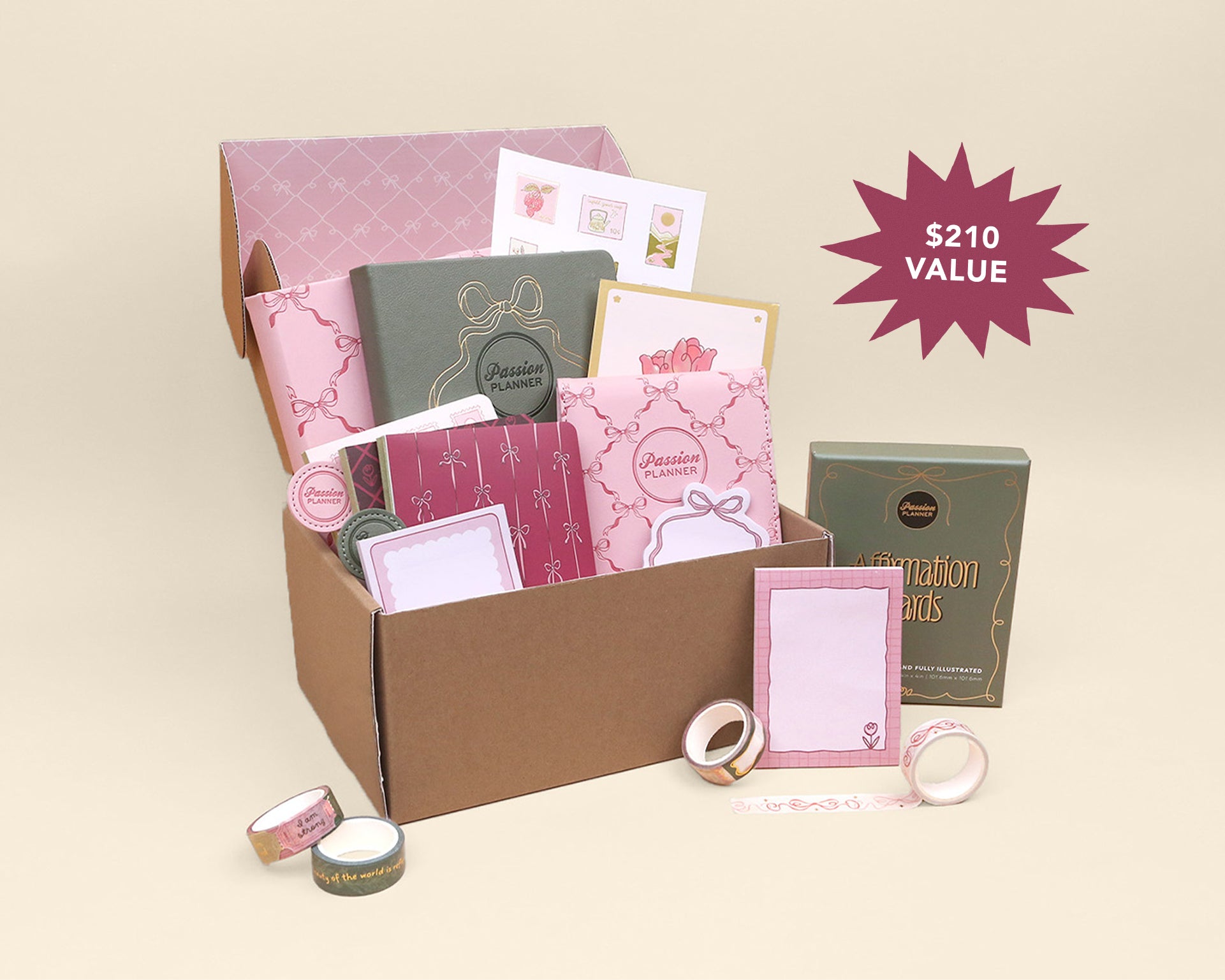 A pink-themed stationery gift set displayed in an open brown cardboard box, with matching boxed items placed beside it on a warm beige background. A magenta starburst in the upper right reads '$210 VALUE'.