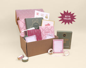 A pink-themed stationery gift set displayed in an open brown cardboard box, with matching boxed items placed beside it on a warm beige background. A magenta starburst in the upper right reads '$210 VALUE'.