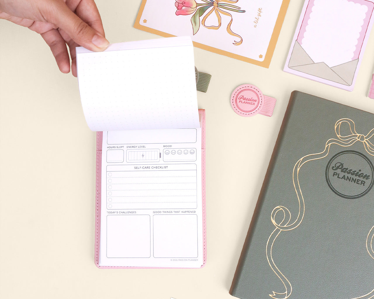 Hand holding a notepad with a visible page, next to a closed planner and stationery items on a light surface.